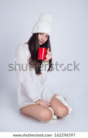 A beautiful brunette woman sitting on the floor in a white sweater and getters no pants a white hat holding a red mug of coffee on a white background