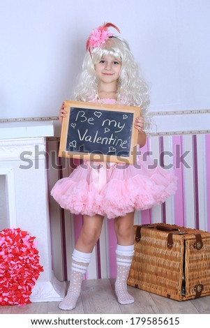A child girl in pink tutu skirt dress holding a chalkboard saying be my valentine in a blond wig