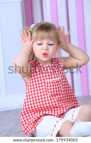 A three year old girl posing like a model in a pink room, in white blouse and pink pants with makeup red lips and a hairstyle making funny face expression