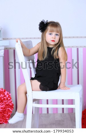 A three year old child girl in a black cocktail dress with a hairstyle and makeup. red lips, sitting on white chair