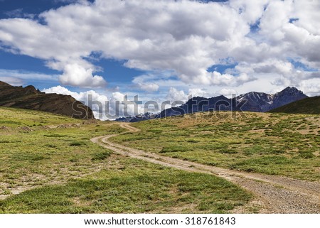Winding dirt road among green meadows going to snow peaks