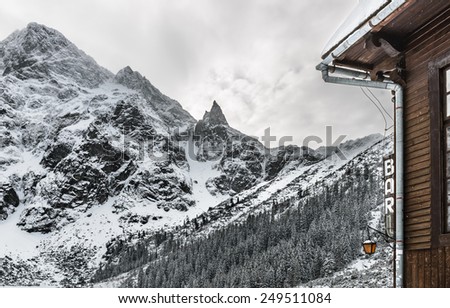 A rustic wooden alpine hut with lamp and bar sign overlooking snow covered mountain peaks on a stormy winter day.