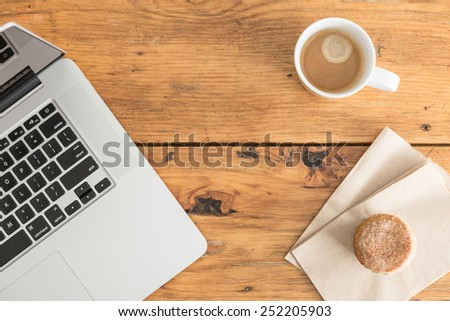 Overhead view of a notebook computer, fresh muffin and a white cup of coffee on a rustic wooden cafe table.