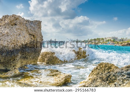 Waves crashing against limestone rocks with the blue Mediterranean Sea and resort hotels in the background on a beach in Paphos, Cyprus.