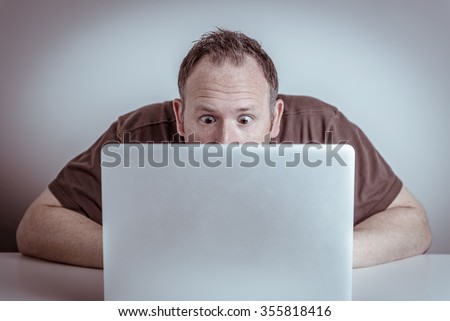 A surprised man with wide eyes sits hidden behind a laptop notebook computer staring at the screen with a shocked or frustrated expression on his face.