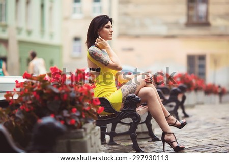 A beautiful woman in a yellow dress, tattoos, sits on the bench, around flowers