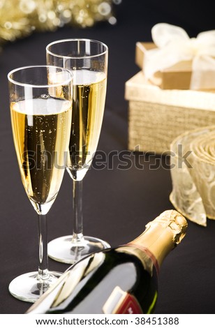 Two flutes and bottle of champagne. Golden gift box and transparent ribbon in background. All on matt black.