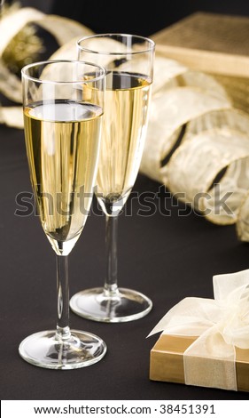 Two champagne flutes, elegant gold gift box with white ribbon. Big gold present and transparent ribbon in background. All on matt black.