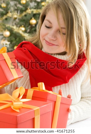 Beautiful, blonde hair, teenage girl looking at presents - red gift boxes with gold ribbon. Christmas tree in background