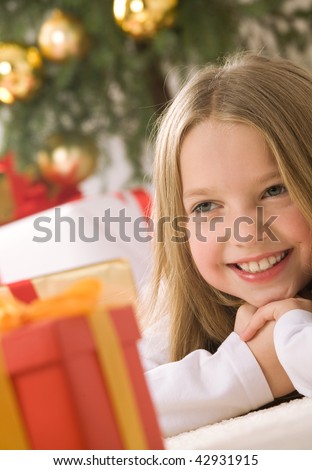 Pretty blond hair girl smiling. Red gift box with gold ribbon on first plan. Christmas tree with golden glass-balls in background