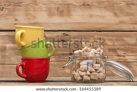 Opened glass jar with sugar with colorful coffee cups on wooden tabletop against grunge wooden background