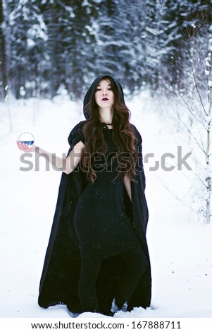 Witch or woman in black cloak with glass ball in white snow forest