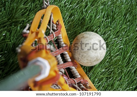 Abstract view of a lacrosse stick scooping up a ball on a green turf field.