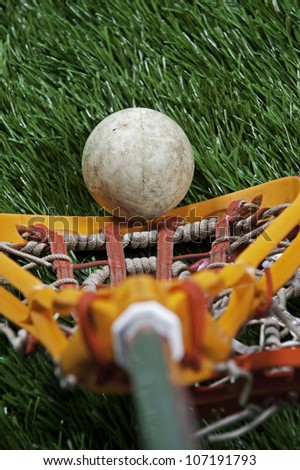 Abstract view of a lacrosse stick scooping up a ball on a green turf field.
