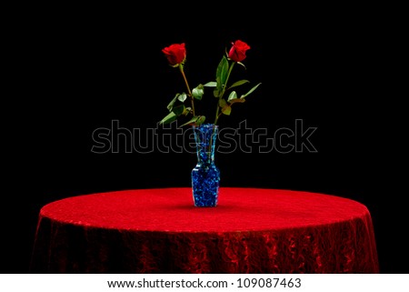 Two red roses in a vase with blue stones on a red lace tablecloth isolated on a black background.