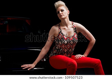 A young sexy woman sits on the bumper of her black car as she waits for assistance in the dark.