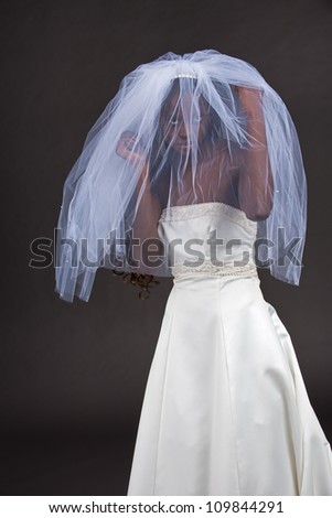 A beautiful young black woman peeks out through the white veil while wearing a beautiful white wedding dress isolated on a grey background.