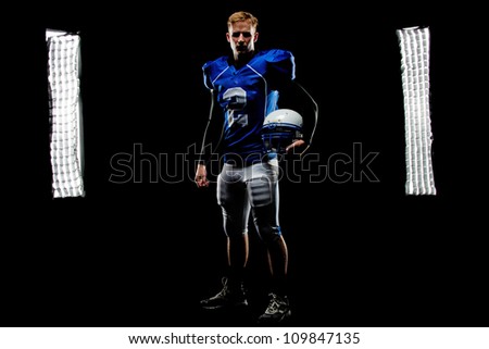 A young football player stands in front of the lights in his uniform holding his helmet.