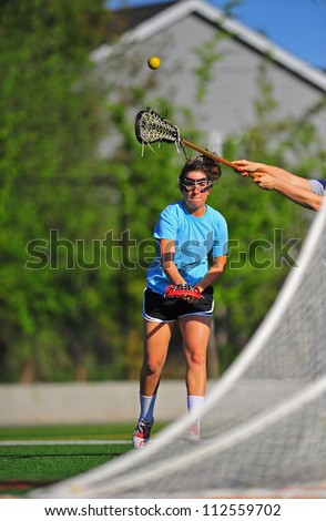 A high school girls lacrosse player takes a shot on goal as a defender stick her stick up to try and block the shot.