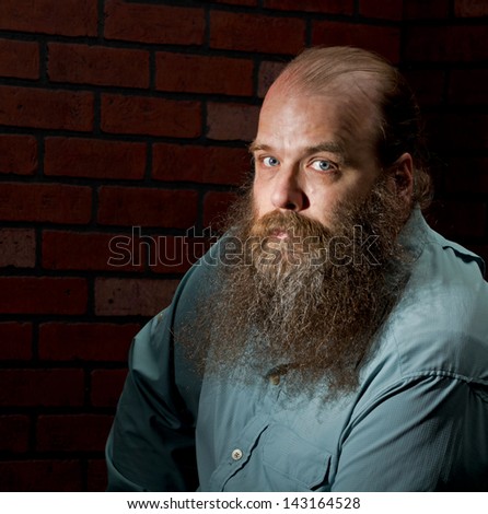 A balding middle aged man with a big bearded against a brick wall looks into the camera with his light blue eyes.