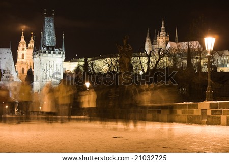The first snow on Charles bridge with walking people at night lighting. Tower bridges, St. Nicholas church and Prague castle in the background.