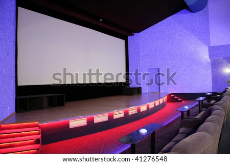 Empty blue cinema auditorium with line of sofa and tables, red stage and projection screen. Side view. Ready for adding your own picture.