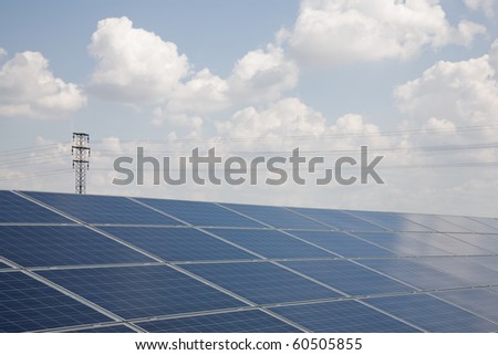 Line of solar power plant panels with clouds and high voltage tower in the background.