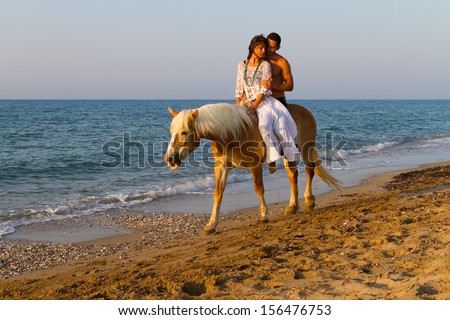 Attractive young couple in love, takes a romantic horse ride along a sandy shoreline in late afternoon summer sun.