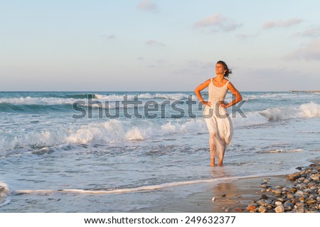 Barefoot young woman in a long white partially wet dress, enjoys a lonesome walk in the water, on a sandy beach in a late summer day, at dusk.