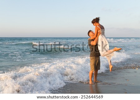 Loving young couple at the beach , in a late summer hazy day at dusk, wearing  a white dress and shorts, enjoying, going barefoot in the ocean water, getting wet, teasing and kissing one another.