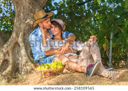 Happy young farmers enjoy a work break sitting at the shade of a tree at the vineyard, eating grapes, kissing, feeding, and teasing one another.