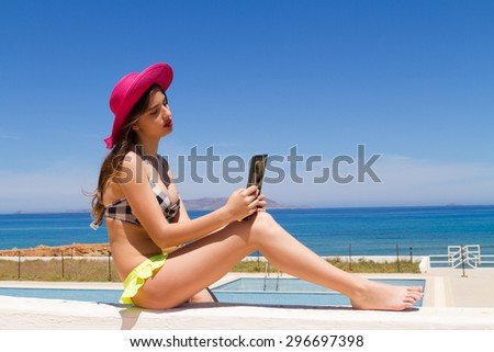 Gorgeous young busty naughty long haired brunette teen girl in swimwear enjoying the sun on a fresh spring morning by the ocean, poses for selfies using her tablet.