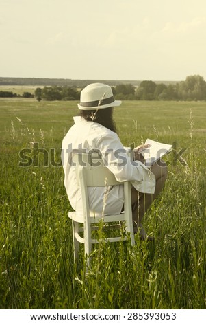 Woman from Behind Sitting on White Chair on Meadow Field and Drawing a Village