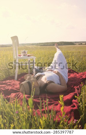 Woman with Hat in White Dress is Laying on Red Cloth on Green Meadow with WHite Chair and Picnic Basket
