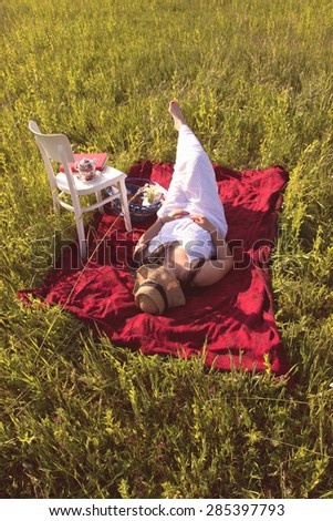 Woman with Hat in White Dress is Laying on Red Cloth on Green Meadow with WHite Chair and Picnic Basket