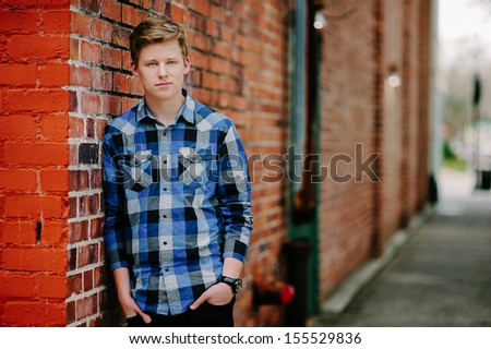 A handsom young man leans against a brick wall in an alley.