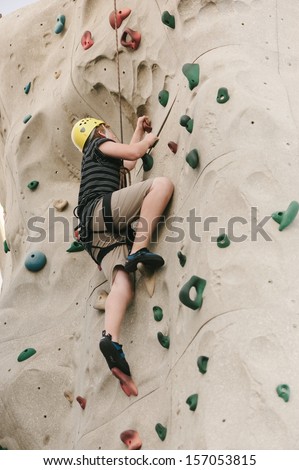 A teen boy climbing on a rock climbing wall with safety harness and helmet on.