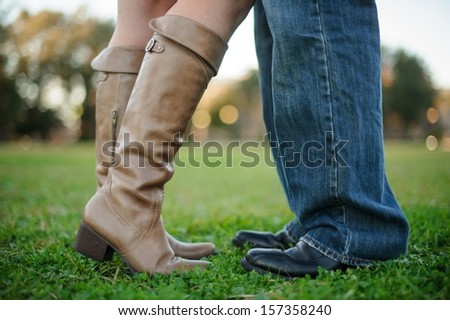 A photo of a couples legs from the knee down where they are on a grassy field.  She has he knee in towards him.