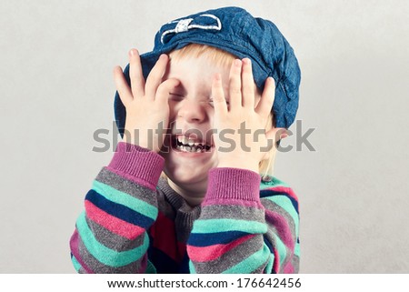 Little girl with a denim cap  covers  face with her  hands, isolated on gray