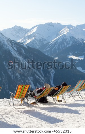 Two skiers relax with a beer in reclining chairs in the sun on top of an Austrian mountain covered with snow