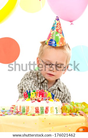A little boy with a birthday cake and balloons in back.