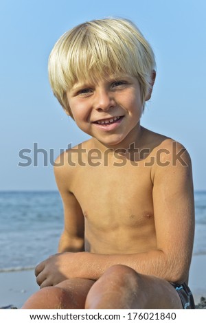 A light haired topless young boy sitting on a beach.