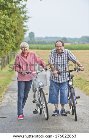 An elderly couple standing next to bikes in countryside.