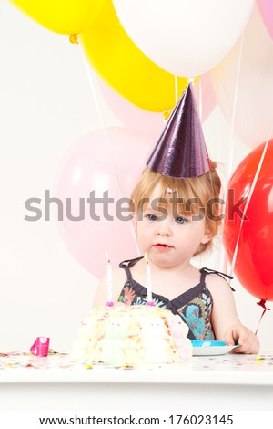 A little girl wearing a purple birthday hat looks at a cake.