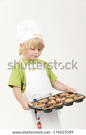 A young boy with apron and chef's hat is holding a tray of muffins.