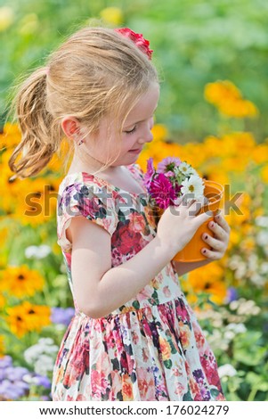 A young girl wearing a flower dress and holding a flower pot full of flowers.
