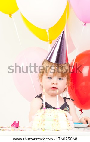 A young girl wearing a purple birthday sitting down with colorful balloons all around her.