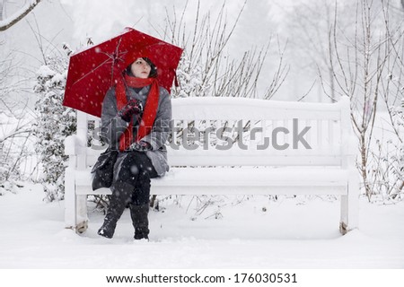 A woman with a red umbrella sitting on a snow covered bench.