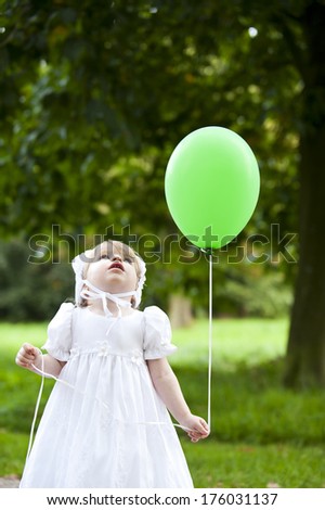A small girl in a white dress has a green balloon.
