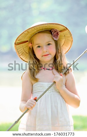 A girl wearing a hat and dress holds a stick.